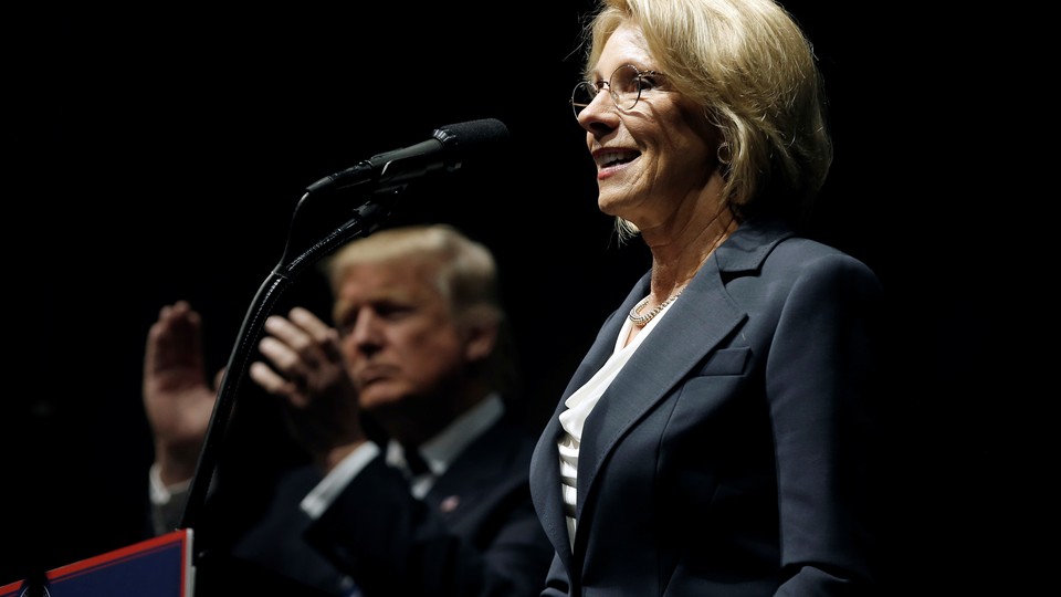 Betsy DeVos stands at a lectern in the foreground of the photo. Donald Trump is clapping behind her.