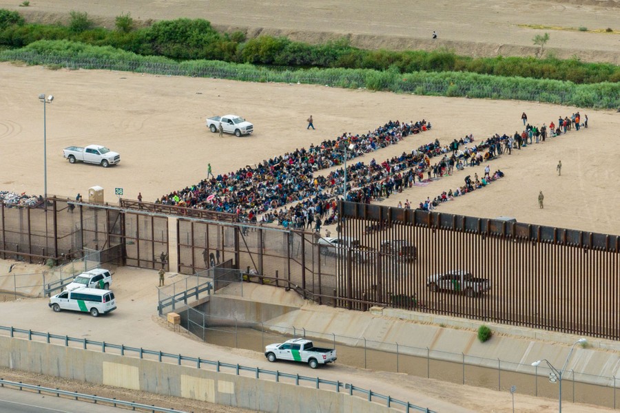 Dozens of people sit on the ground in several lines beside a tall border fence.