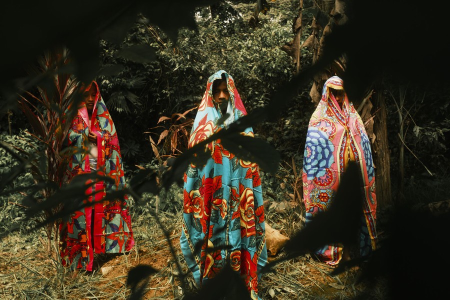 Several young people stand among trees, each draped in a colorful patterned cloth.