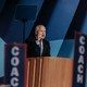A man in a suit and blue tie with balding white hair speaks from a podium with signs that say coach around him.