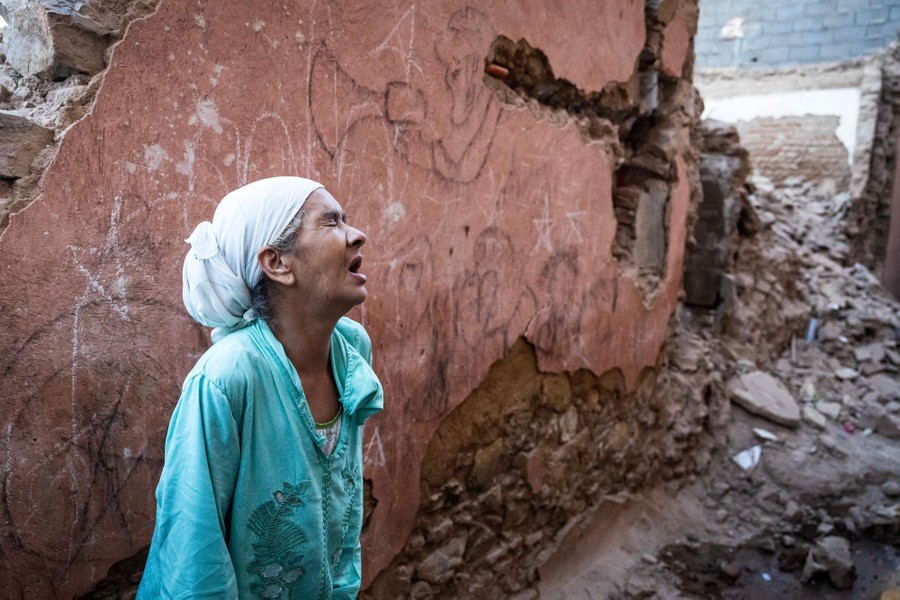 A woman stands with her eyes closed and mouth open, as if in anguish, in front of a damaged wall and piles of rubble, after an earthquake.