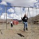 Two children play on the swings outside a red schoolhouse. The setting is rural.