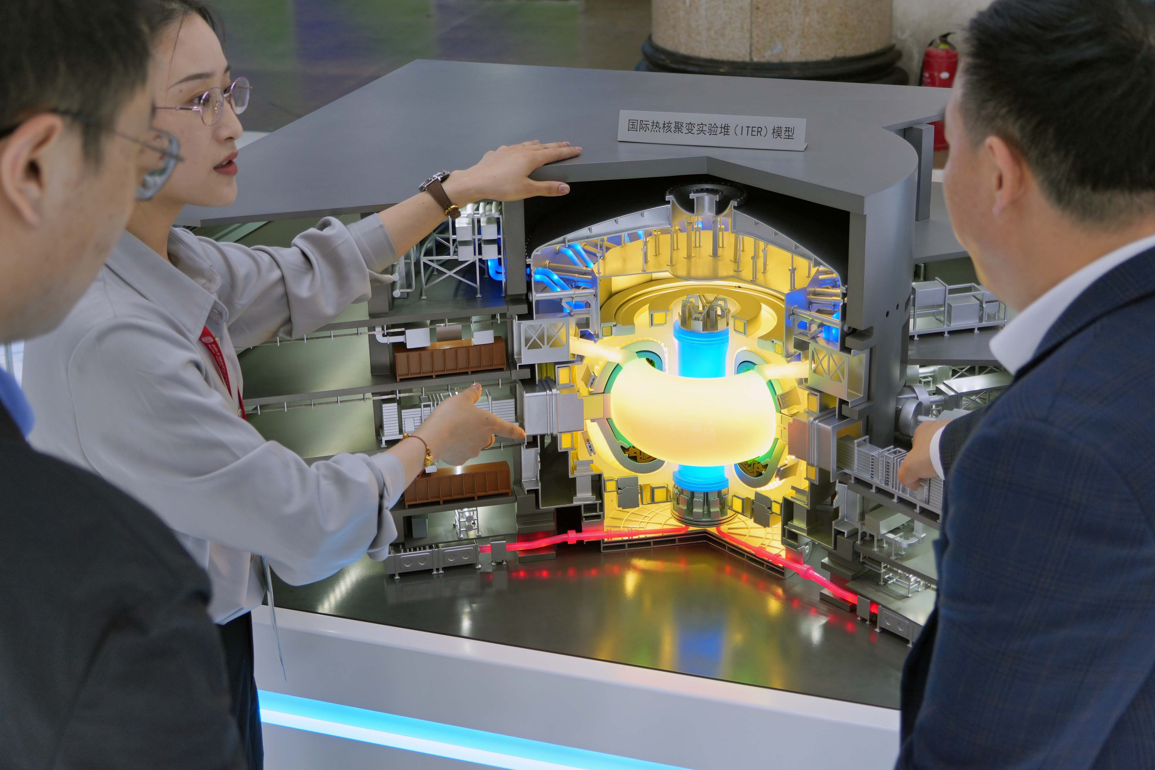 Several people gesture toward and look at a scale model depicting a nuclear-fusion reactor.