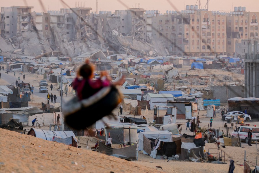 A child plays on a tire swing near a tent city and a number of damaged and destroyed residential buildings.
