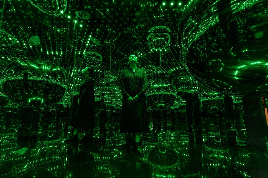 A person poses inside a room with walls, ceiling, and floor all covered by mirrors, illuminated by many small green lights.