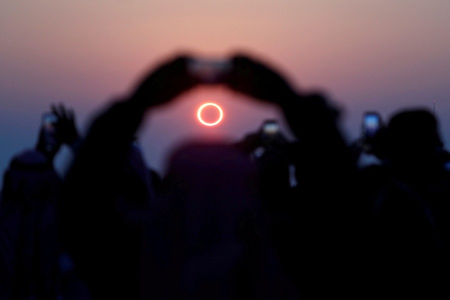 Several people hold up their phones to take pictures of an eclipse, the distant sun appearing like a bright ring, partly blocked by the passing moon.