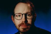 Ari Aster wearing glasses against a blue background