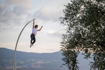 A performer hangs from the end of a tall curved pole, by one hand.