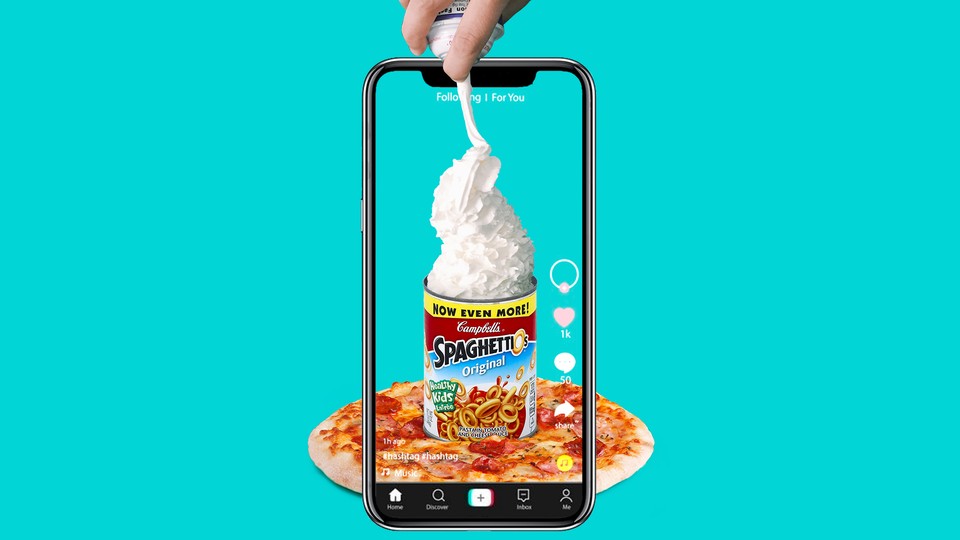 A phone records a hand spraying whipped cream into a can of Spaghetti-Os on top of a pizza.
