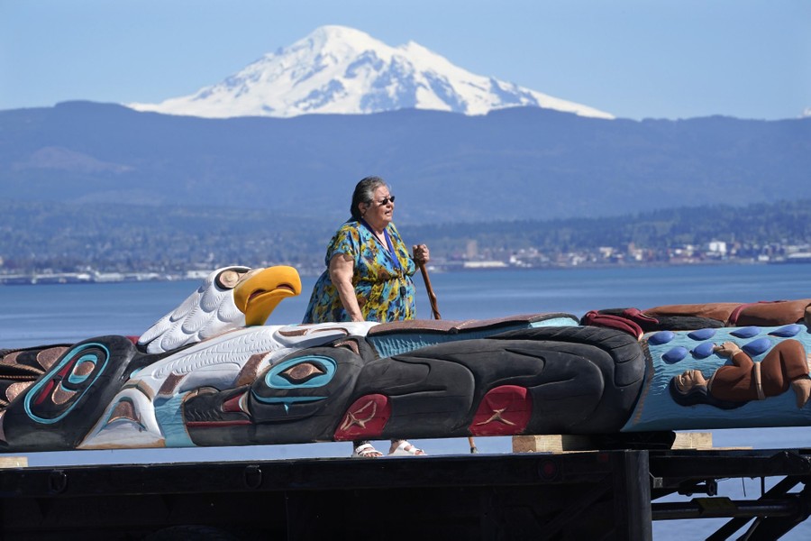 A woman walks past a large totem pole that is lying down; a snow-capped mountain and a harbor are visible in the background.