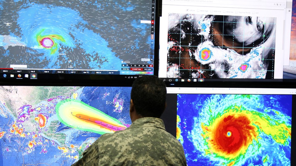 A man in camouflage stands in front of computer screens showing images of Hurricane Irma. 