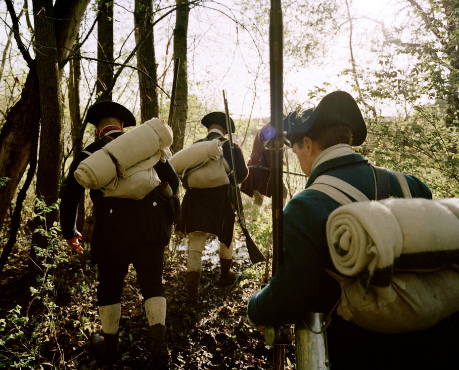 photo of reenactors as American soldiers wearing packs with bed rolls and hiking in a line through the woods towards Fort Ticonderoga