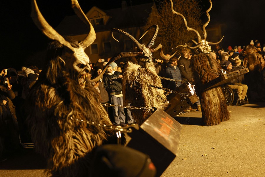 Several people dressed in large frightening Krampus costumes carry heavy metal drums while marching in a parade.