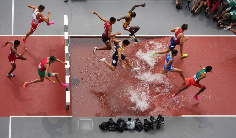 An elevated view of runners jumping over a hurdle and splashing in shallow water during a race.