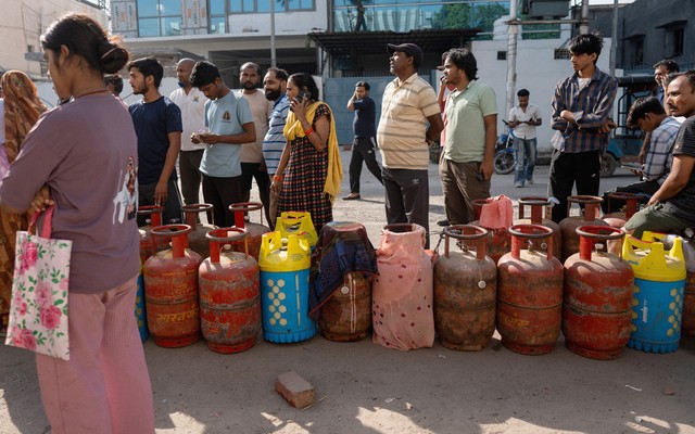 Indians wait in a long line beside red, pink, and blue gas cylinders. Some are talking on the phone, while others stare ahead with their arms crossed.