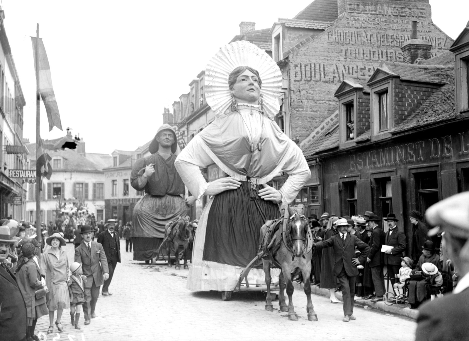 Horse-drawn carts carry tall figurines down a street during a parade.