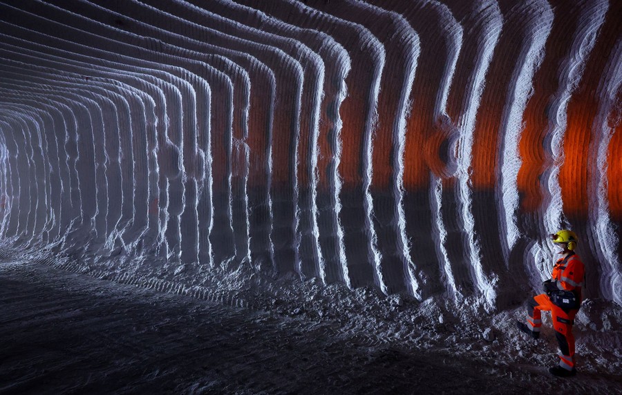 A miner stands inside a large underground tunnel, with parallel ridges along its walls and ceiling left by digging equipment.