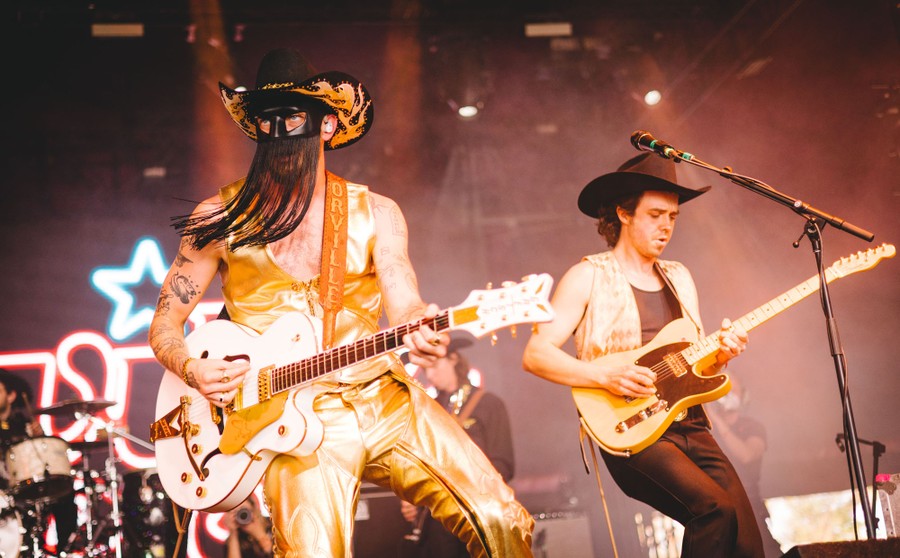 Two performers play guitars onstage. The performer closest to the camera wears a cowboy hat and a mask over their eyes with long fringes hanging from it over their lower face.