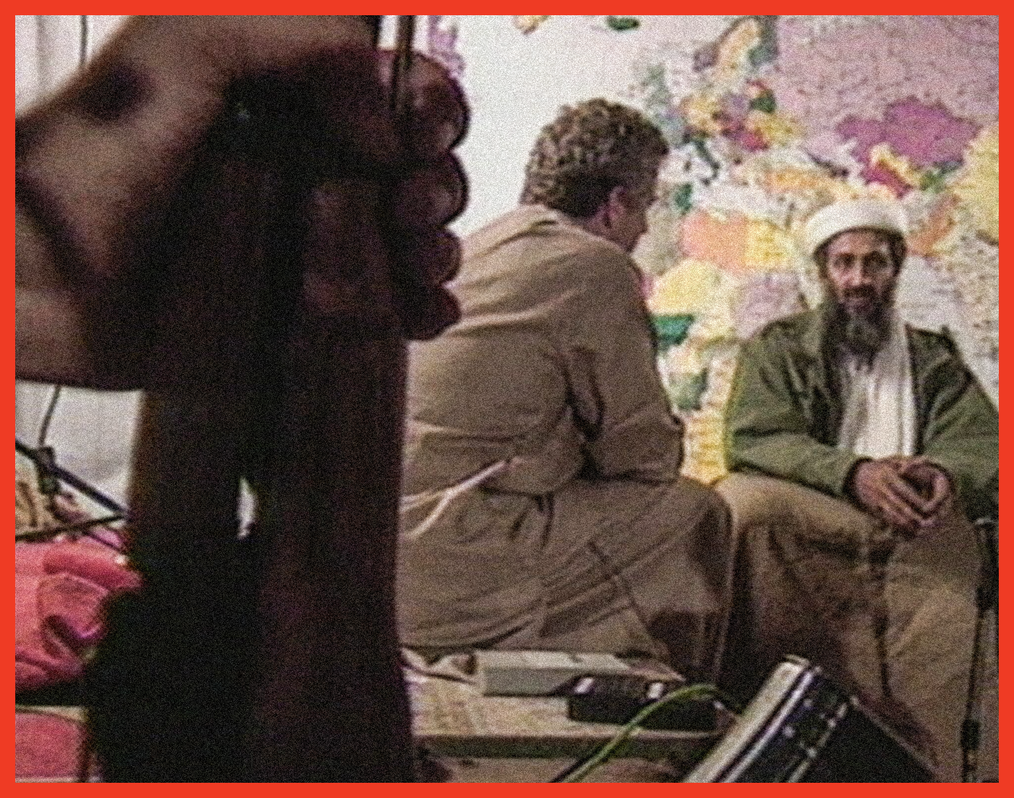 video still from ABC interview of hand holding rifle in foreground and man in background interviewing bin-Laden seated in front of world map