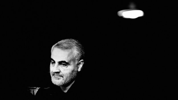 A black-and-white photo of Qassem Soleimani, looking downward to the left of the camera