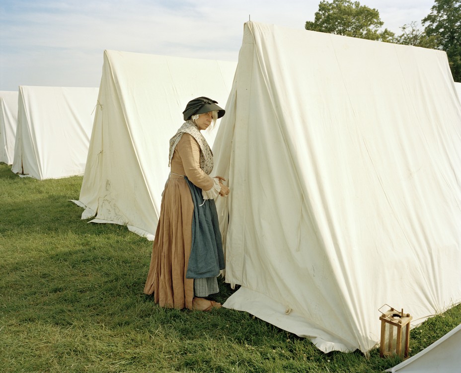 photo of woman in colonial dress, apron, and hat looking sideways at viewer as she enters one of a row of white-cloth triangular tents pitched on the grass at Gloucester, MA during a reenactment of the Battle of Bunker Hill
