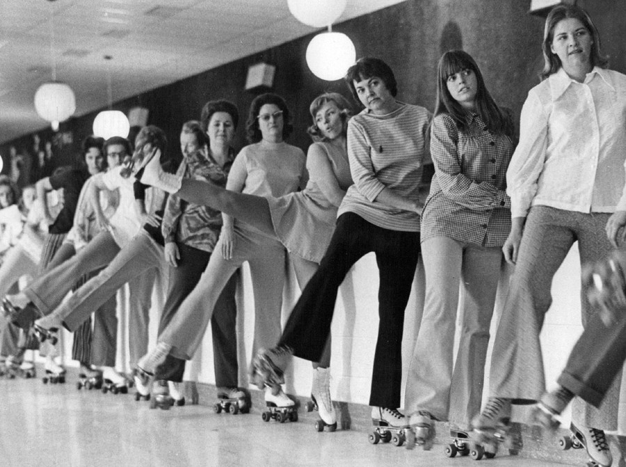 A line of people wearing roller skates lift one leg while leaning against a wall.