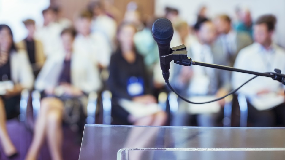 Close-up of microphone and transparent lectern with audience seen in blurred background