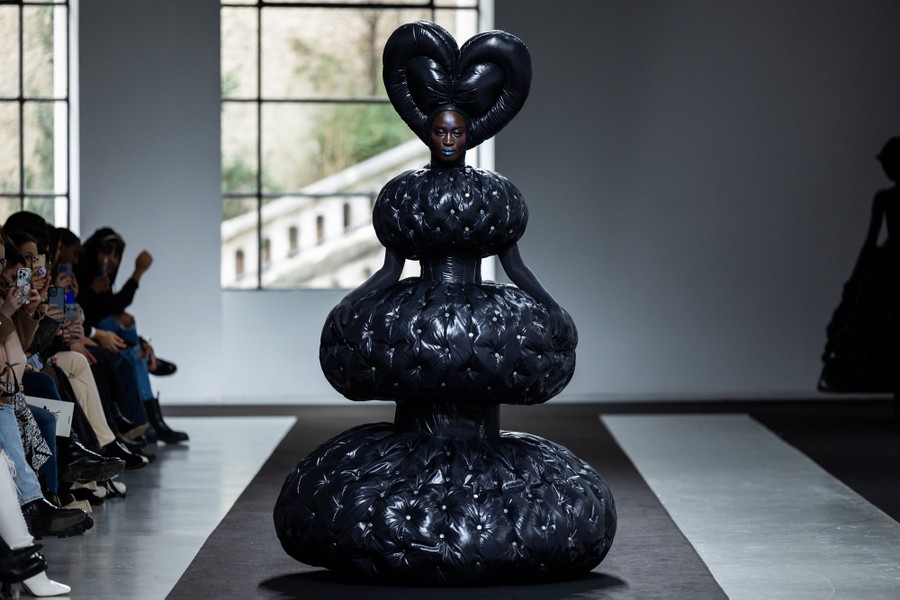 A model on a runway wears a heart-shaped headdress and an elaborate, puffy garment that together suggest a black chess piece.