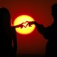 Two young people hold their hands close together, their fingers forming a heart shape in silhouette against the setting sun.