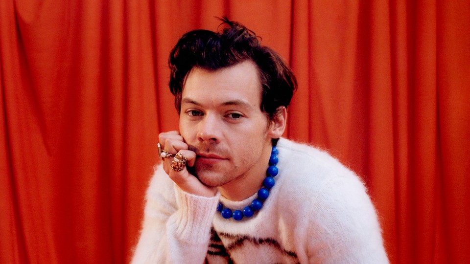 Harry Styles in a white sweater, blue chunky necklace, against orange-red curtain backdrop