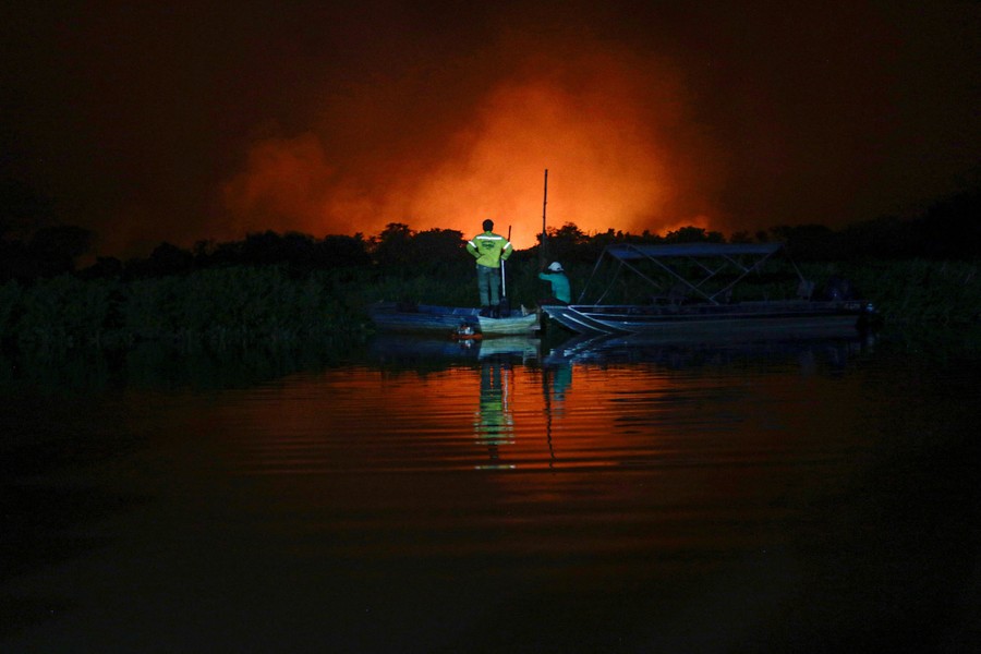 Two people watch the glow of a distant wildfire from a riverbank, at night.