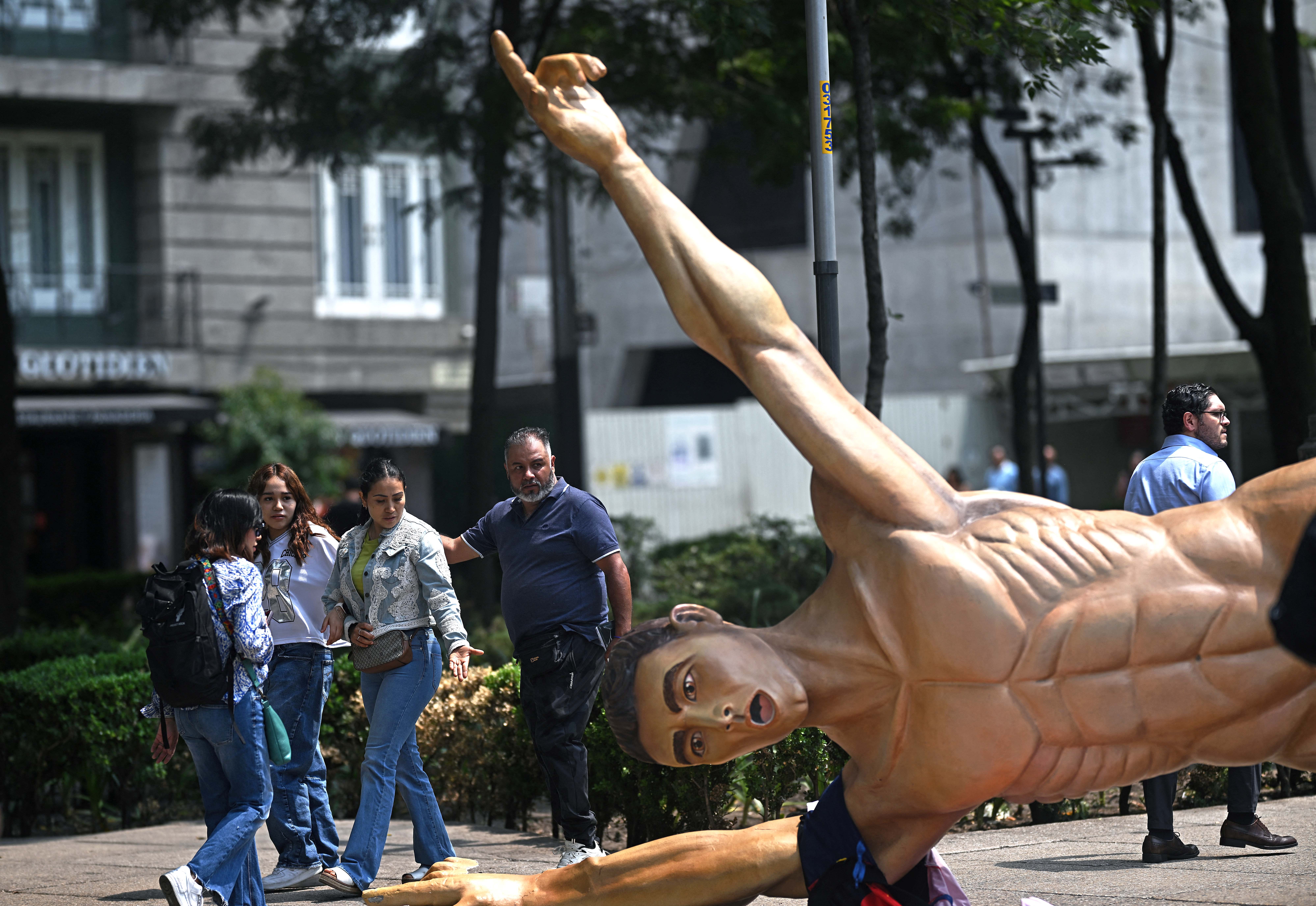 People walk past a toppled sculpture of a celebrating soccer player.