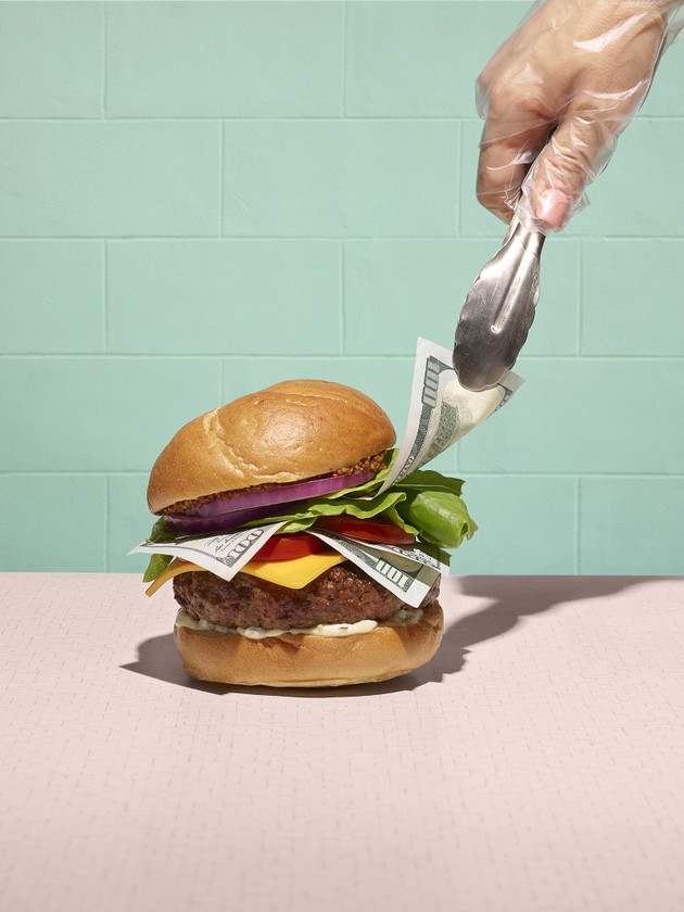 Gloved hand uses tongs to pry dollar bills from a burger stuffed with cash.