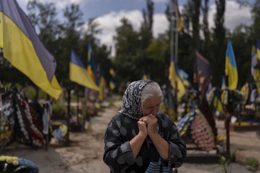 A woman holds her hands to her face, crying, in a cemetery, surrounded by Ukrainian flags.
