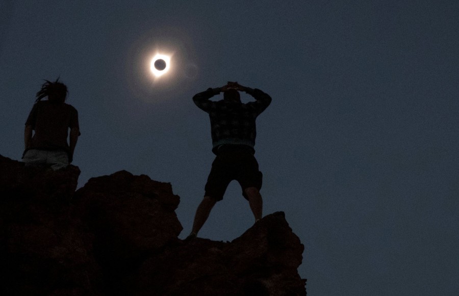 Two people stand on a rocky outcrop, looking up toward the eclipsed sun.