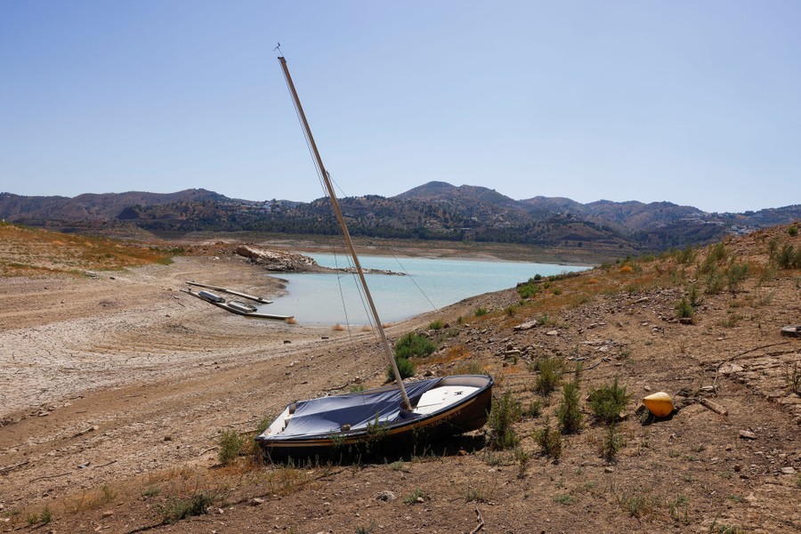 A small sailboat sits on the dry sloped shore of a reservoir with very low water.