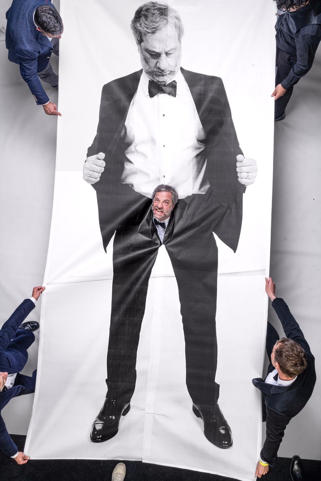 photo from above of 4 blue-coated men holding the corners of a large black-and-white banner printed with photo of Apatow in a tuxedo holding open his jacket and looking down at his fly, where the real Apatow has pushed his face through the banner's ripped fabric
