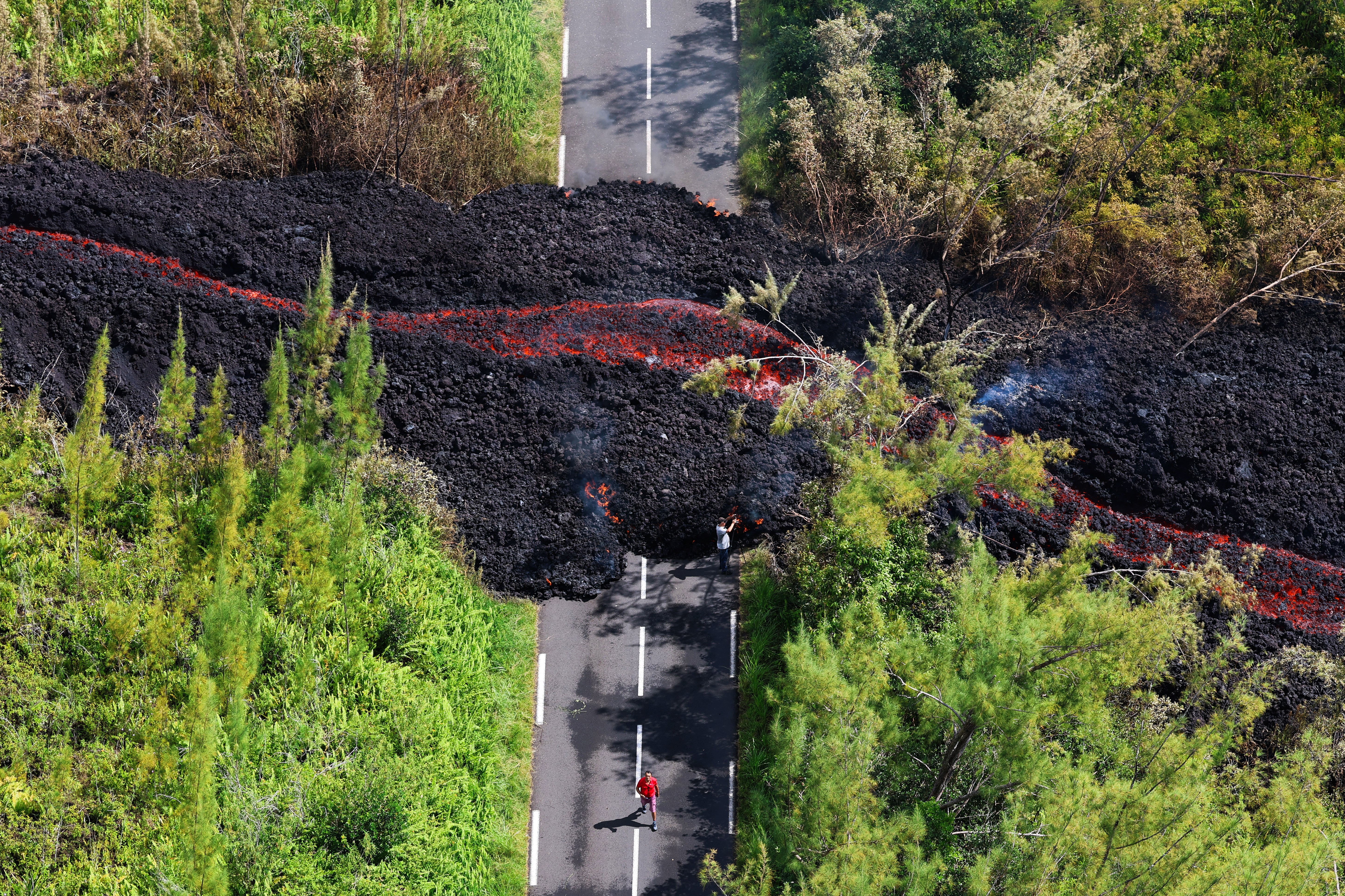An aerial photograph shows a lava flow cutting across a paved road in a forested area.