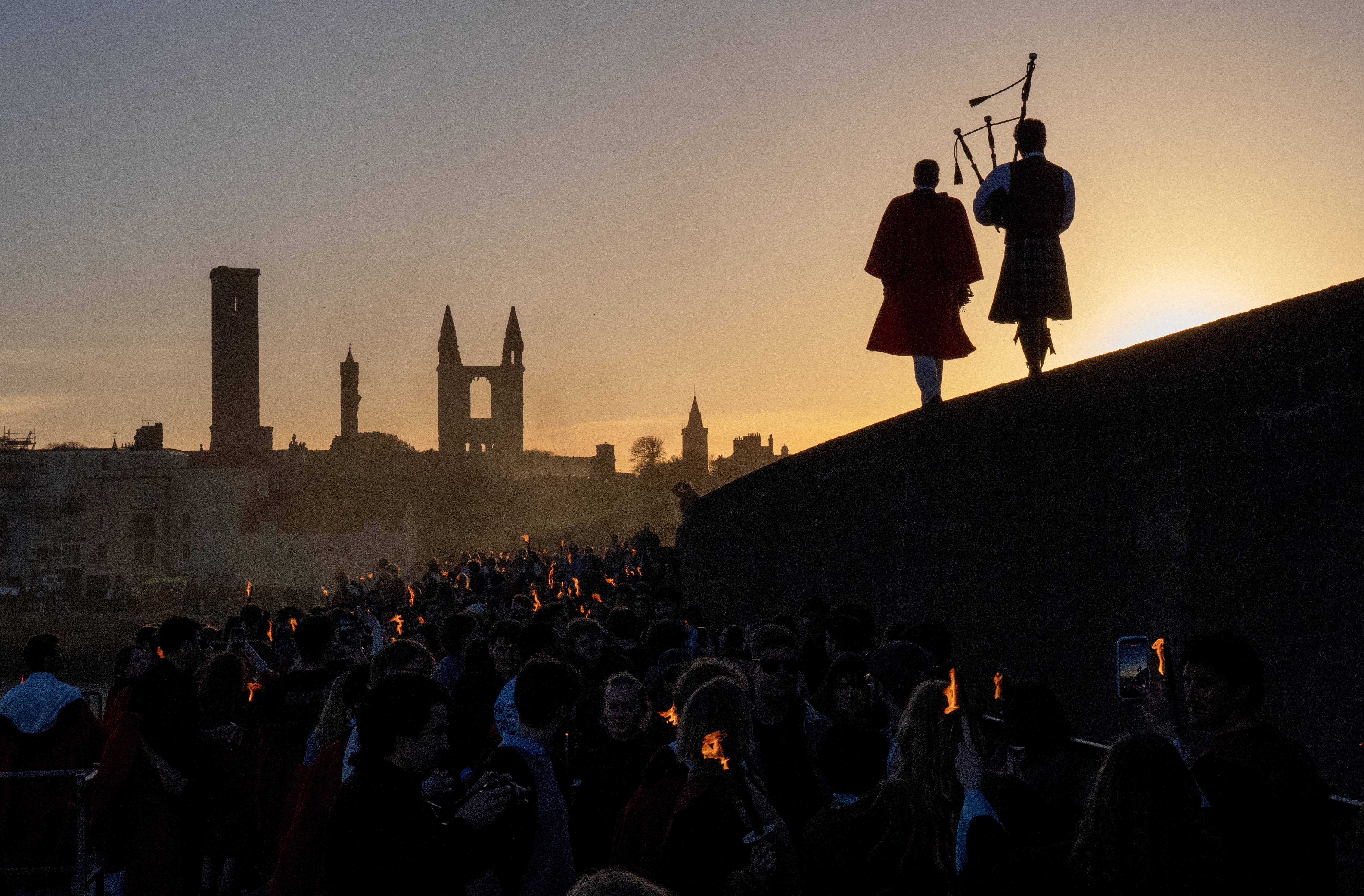 Two people, one with a bagpipe, walk on a wall, leading a procession below them toward a pier.