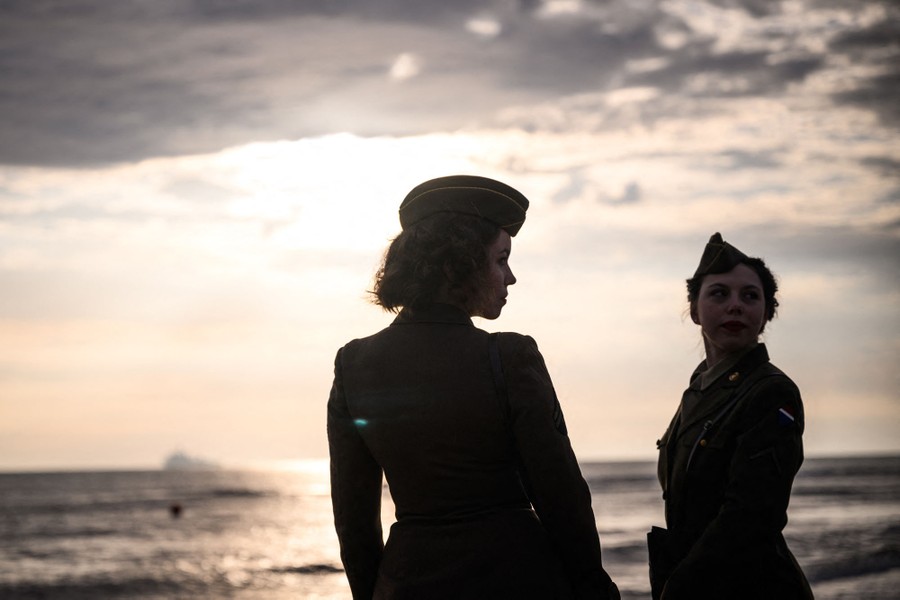 Two people in WWII-era military attire stand on a shoreline.