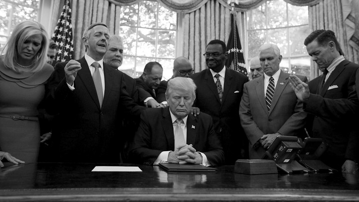 President Trump, Vice President Mike Pence, and faith leaders say a prayer in the Oval Office in September 2017. Several people have their hands on Trump's shoulders. Most of the people have their eyes closed.