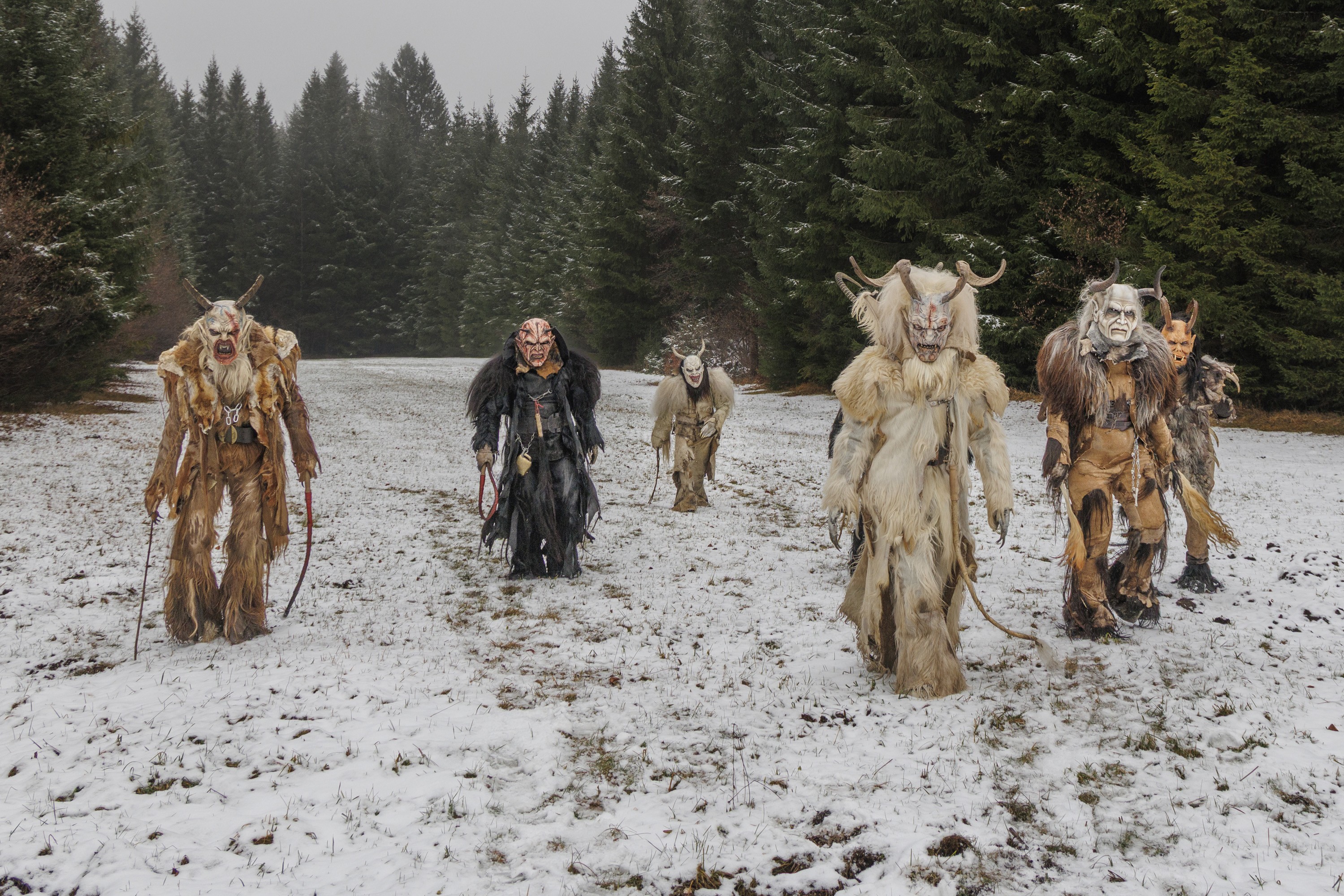 Six performers wearing frightening Krampus costumes and masks walk across a snow-covered field out of a forest.