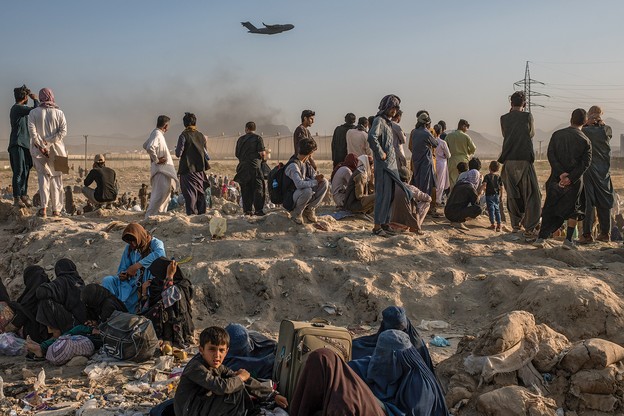 Women and children sit on dusty rocks with men standing behind, facing distant airport walls and watching a military plane taking off