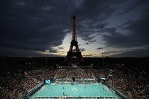 A view of a small football stadium at night, filled with spectators, beneath the Eiffel Tower