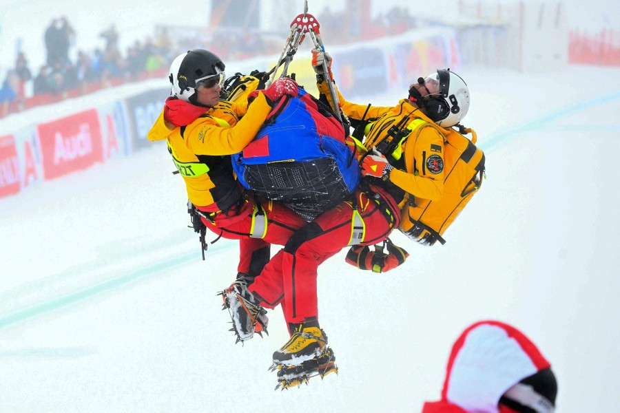 Two rescuers hang alongside a stretcher carrying an injured skier beneath a helicopter above a ski run.