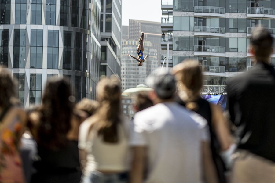 A crowd watches a high diver with city buildings visible in the background.