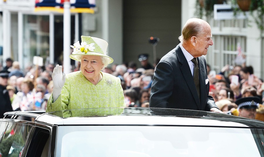 The Queen and her husband wave to onlookers while standing in an open-top car.