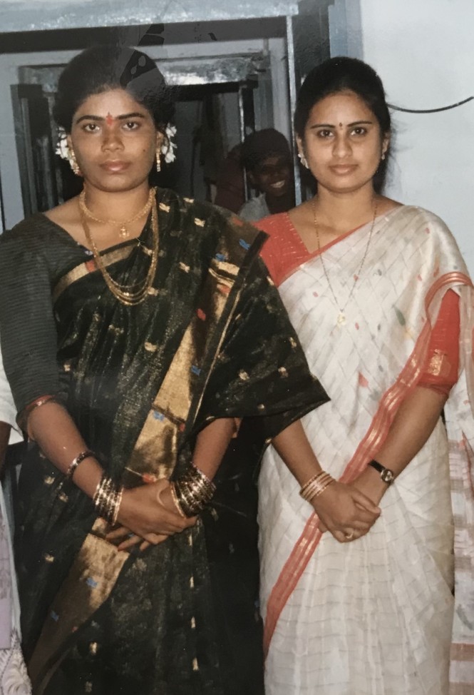 Two women wearing saris stand with their hands clasped