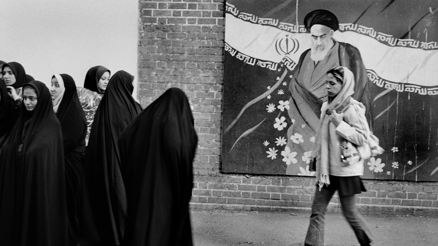 a woman in a headscarf walks in front of a mural showing Ruhollah Khomeini and toward a group of women in black chadors