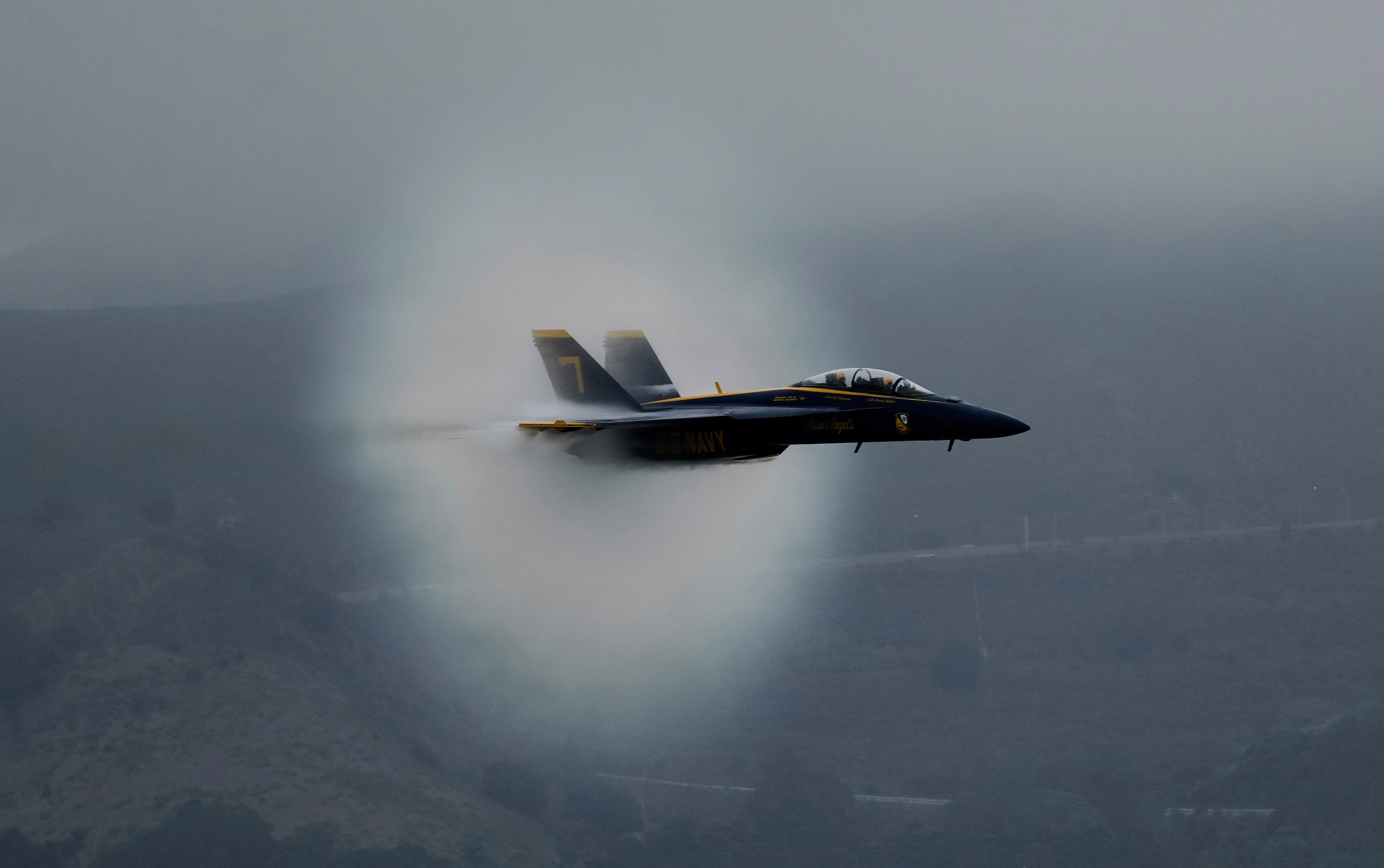 A Blue Angels jet aircraft flies past, generating a cone-shaped condensation cloud.